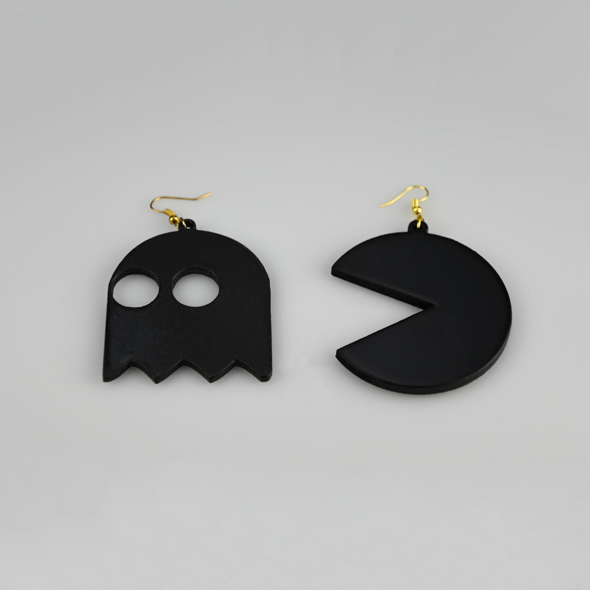 A pair of black dangling earrings shaped like a ghost and a Pac-Man style circle, each with a gold fishhook ear wire, photographed on a plain light gray background.