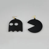 A pair of black dangling earrings shaped like a ghost and a Pac-Man style circle, each with a gold fishhook ear wire, photographed on a plain light gray background.