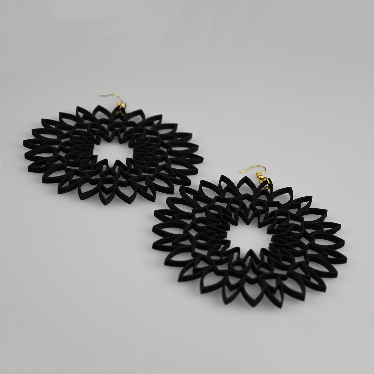 A pair of black laser-cut dangle earrings with a geometric, sunburst-like pattern and small gold fishhook wires, displayed on a light gray background.